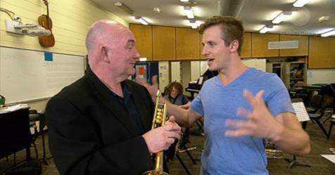 Two people talking inside a conference hall, with one of them holding a trumpet, and the other guy in blue tshirt